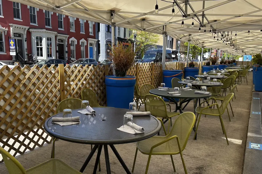 A shaded outdoor dining setup features neatly set green chairs and tables under a canopy along a vibrant urban street.