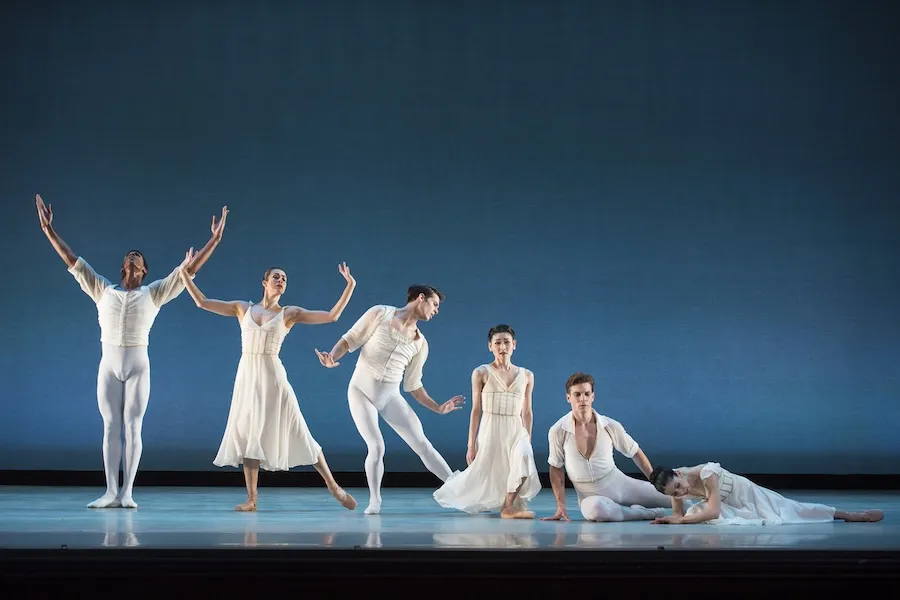 Six ballet dancers in white costumes perform a graceful contemporary piece on a minimal stage.