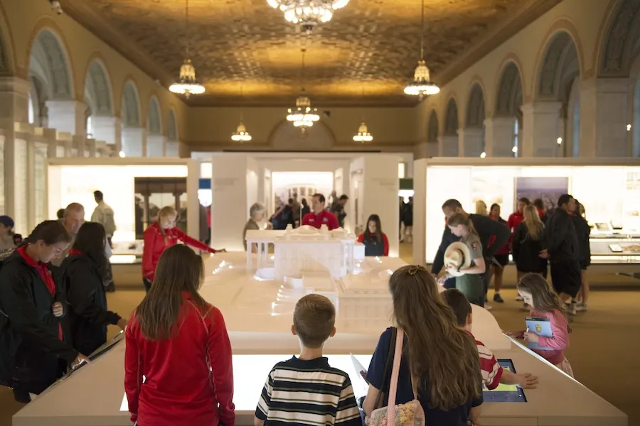 Visitors of all ages explore a scale model of the White House inside a grand exhibition hall.
