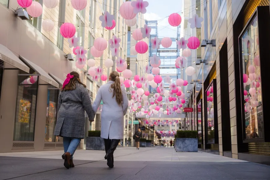 A couple in winter coats walks through modern storefronts with pink lanterns hanging above. 