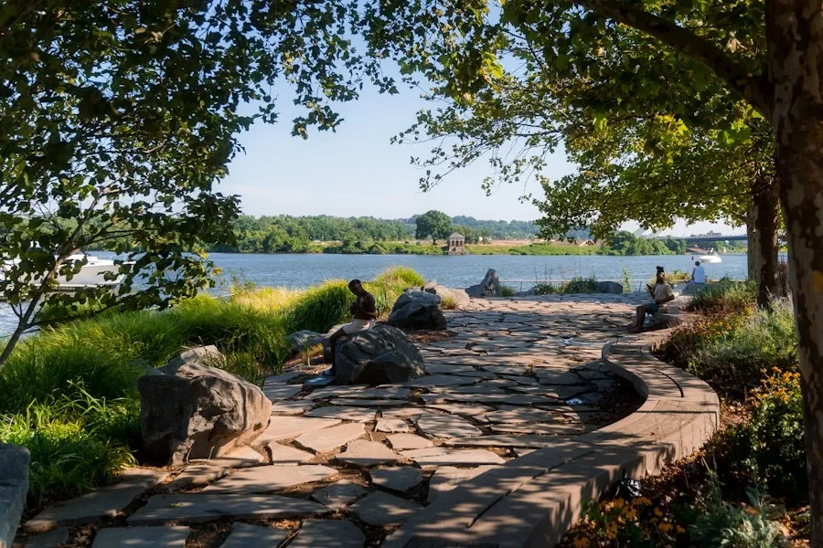 A stone pathway curves along the Anacostia River with people relaxing under shady trees at Yards Park.