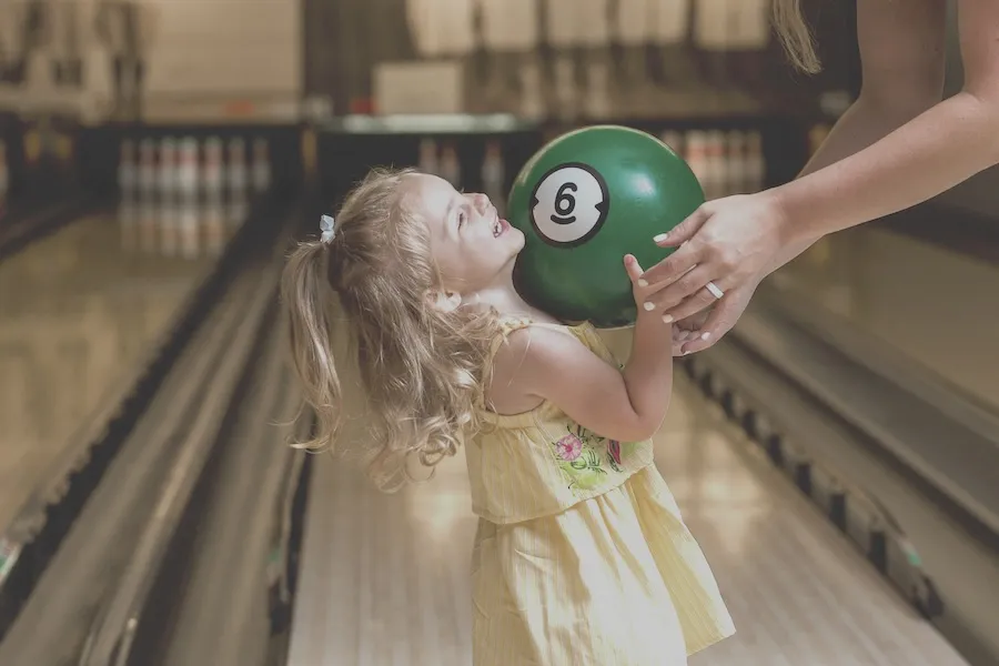 A smiling child holds a bowling ball in a bowling alley, with an adult reaching out hands to help. 