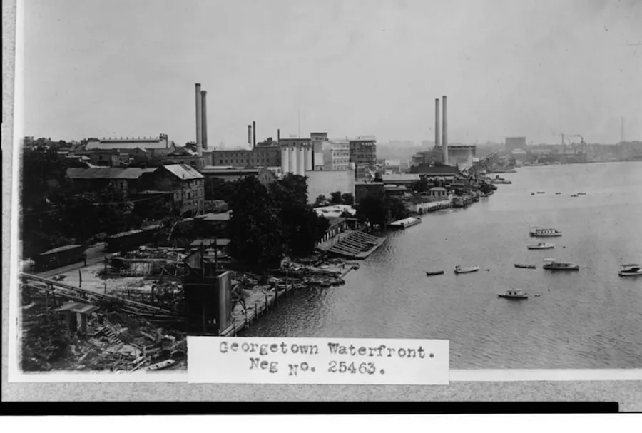 lack-and-white photo of the historic Georgetown waterfront with industrial buildings, smokestacks and boats on the Potomac River.