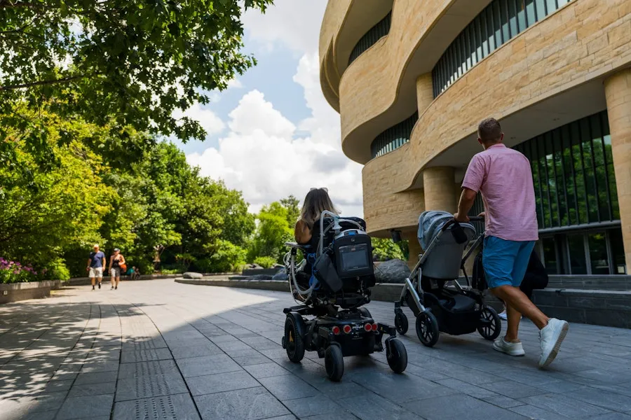 A person using a motorized wheelchair and a man pushing a stroller outside the National Museum of the American Indian.
