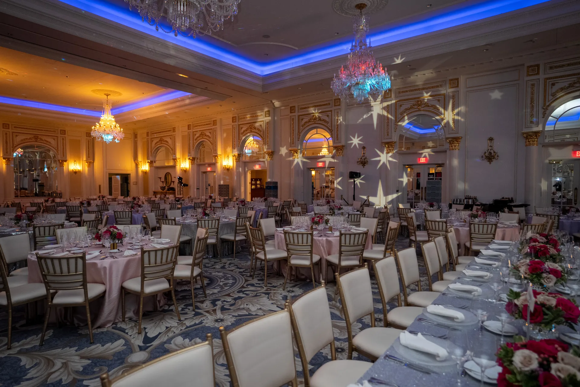 Un'elegante sala da ballo allestita per una cena formale, con lampadari, proiezioni luminose a forma di stella, tavoli rotondi con tovaglie rosa e centrotavola floreali.