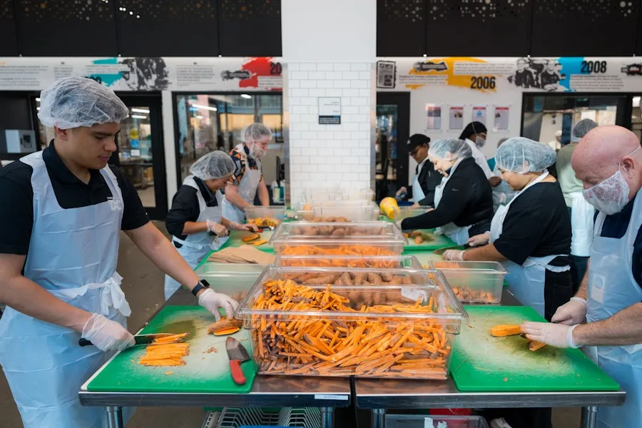 Volunteers wearing aprons and hairnets chop and prepare carrots together at a community kitchen.