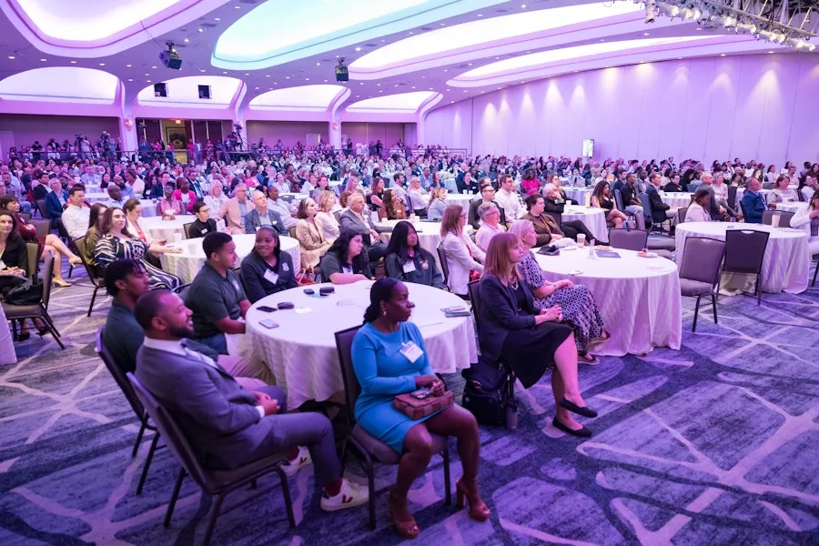 A large audience sits at round tables in a purple-lit conference hall, attentively listening to a presentation.