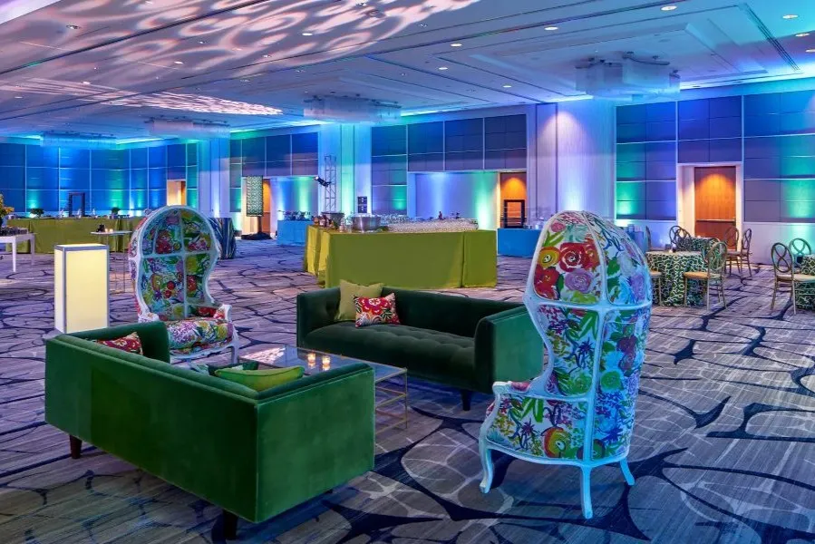 Un colorato allestimento per eventi in sala da ballo, con divani in velluto verde, sedie con schienale alto e motivi floreali, tavoli da cocktail e un'illuminazione d'accento vivace nei toni del blu e del verde.