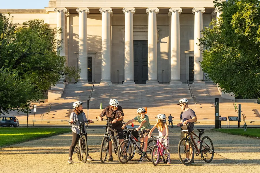 A group of people with bicycles, including two children, stop in front of the National Gallery of Art in Washington, DC, during a guided bike tour.