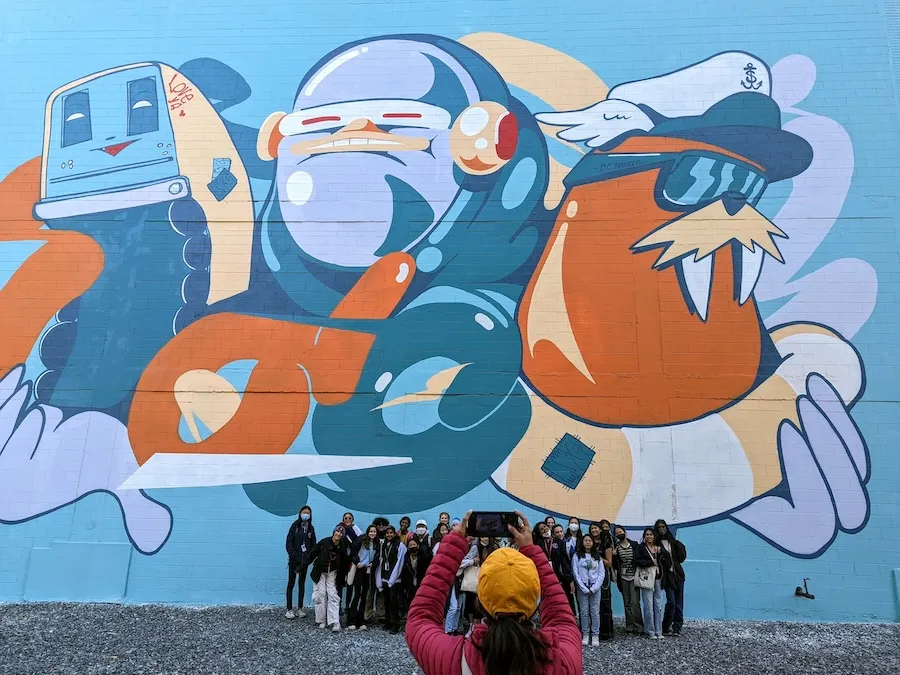 A group stands in front of a large colorful mural in Washington, DC, as one person takes a group photo.