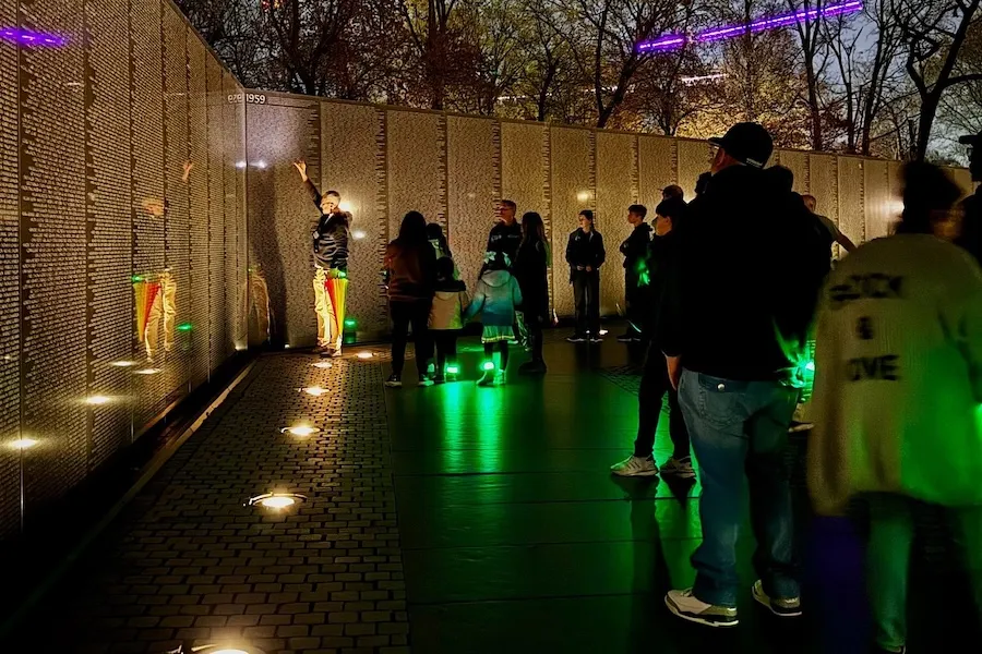 A group of travelers listen to a tour guide discuss the Vietnam Veterans Memorial in DC at night, illuminated by LED bracelets. 