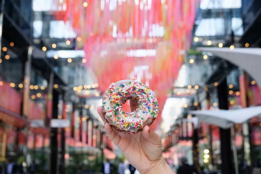 A hand holds up a sprinkle-covered donut in front of colorful streamers hanging above Palmer Alley in CityCenterDC.