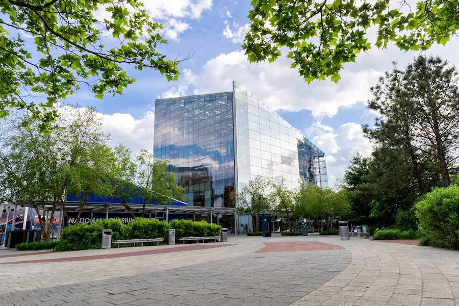 A view of the glass-front National Aquarium building surrounded by trees and greenery under a partly cloudy sky in Washington, DC.