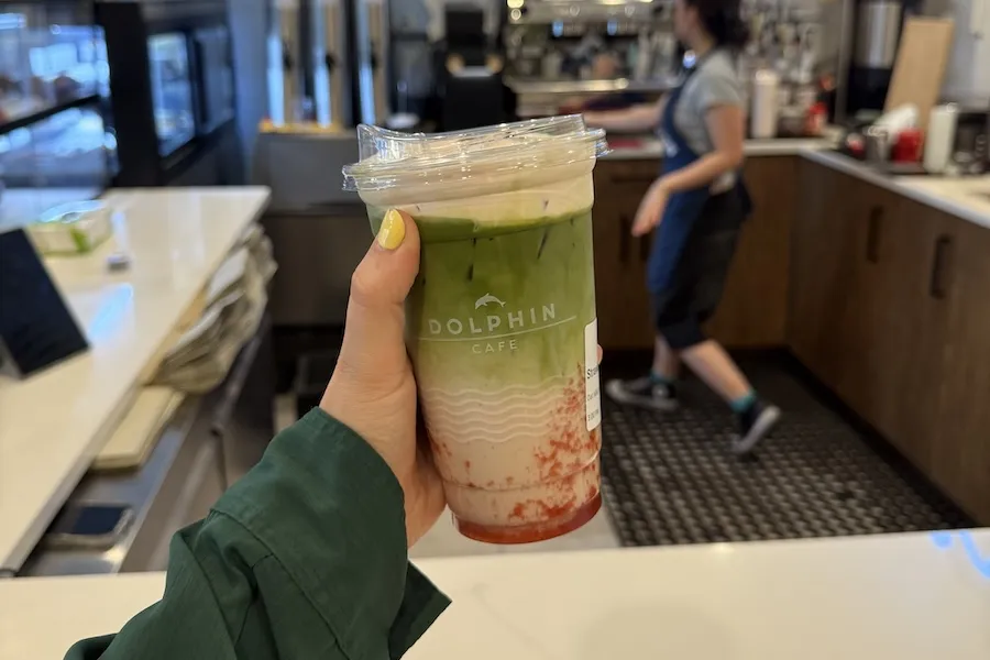 A hand holds out a matcha drink with a splash of pink at the bottom of the cup. 