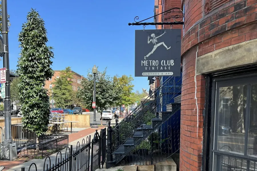 The sign for Metro Club Vintage, a shop selling clothes and art, hangs outside a brick building on a sunny street.