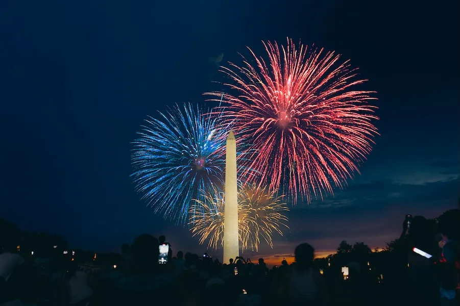 Fireworks burst in red and blue above the Washington Monument as crowds watch on the National Mall at night.