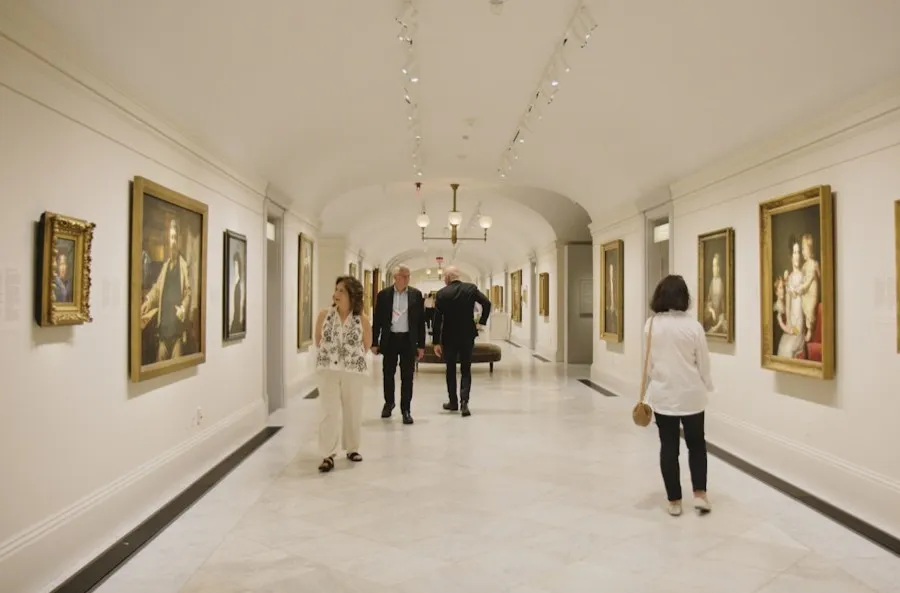 Visitors walk through a long, elegant museum gallery lined with framed paintings on white walls under soft, evenly spaced lighting.