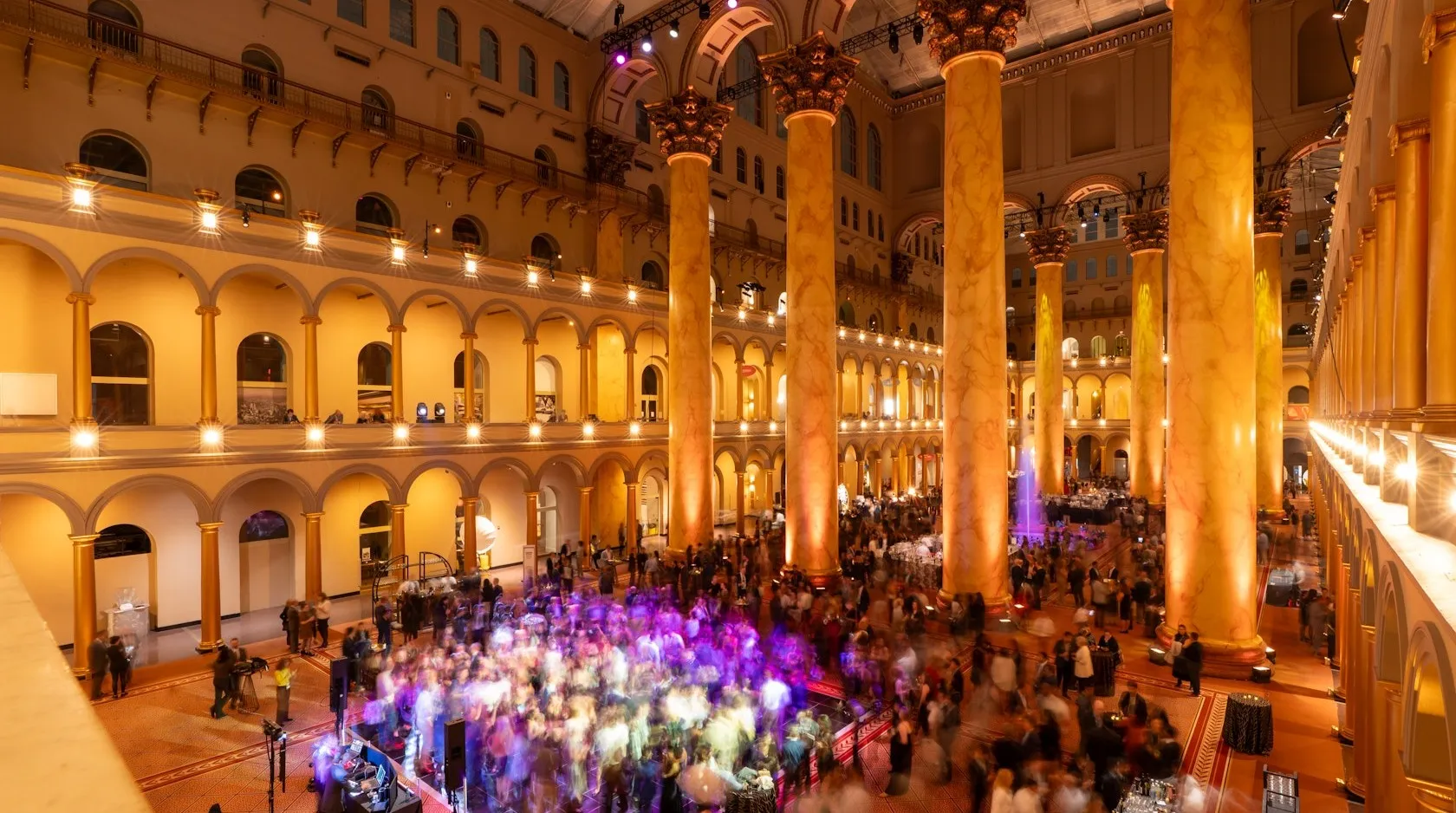 A large crowd mingles beneath towering golden columns and arched balconies inside a grand, brightly lit hall during a lively evening event.