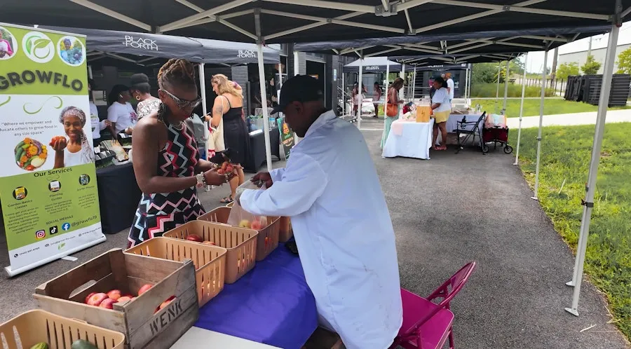 A vendor and customer exchange produce beneath tents at a neighborhood farmers market.