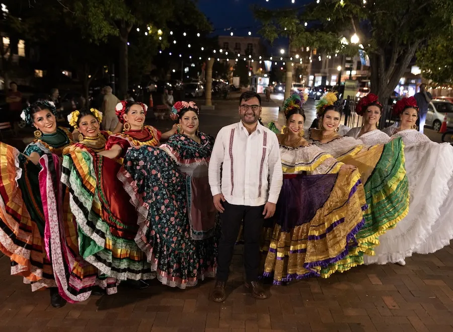 Performers in colorful traditional dresses pose with a man on a lively city street at night.