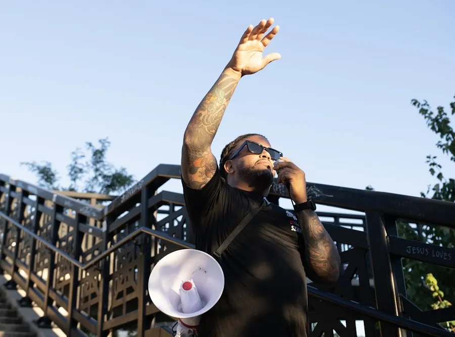 A run club leader holds a megaphone and raises his hand while addressing a group outdoors.