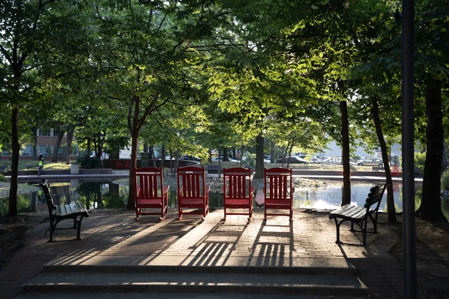 Red chairs line a shaded platform beside a calm pond in a leafy park.