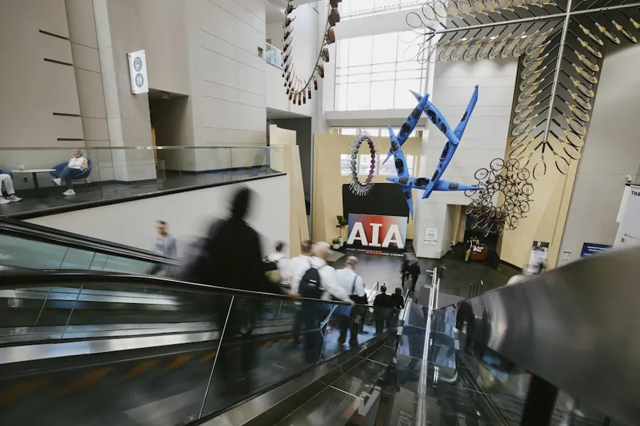 Visitors descend an escalator into a bright museum atrium featuring suspended sculptural installations and a prominent AIA display.