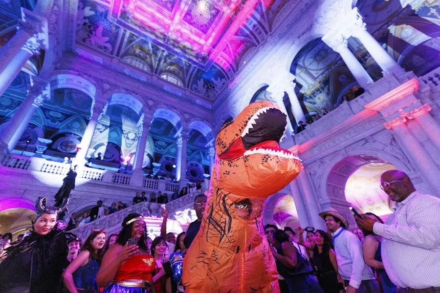 A person in an inflatable T-rex costume dances among a crowd at a colorful, neon-lit event inside an ornate historic hall.