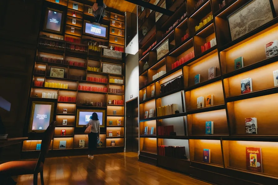 A person explores a warmly lit, floor-to-ceiling bookshelf display with interactive screens in a modern library-like space.