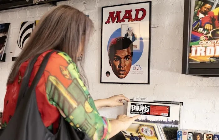 A woman flips through vinyl records in a shop beneath framed posters on the wall.