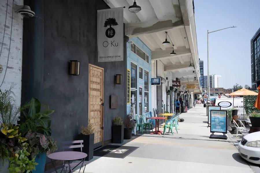 A row of storefronts features outdoor seating and signage outside a restaurant along a sunny sidewalk.