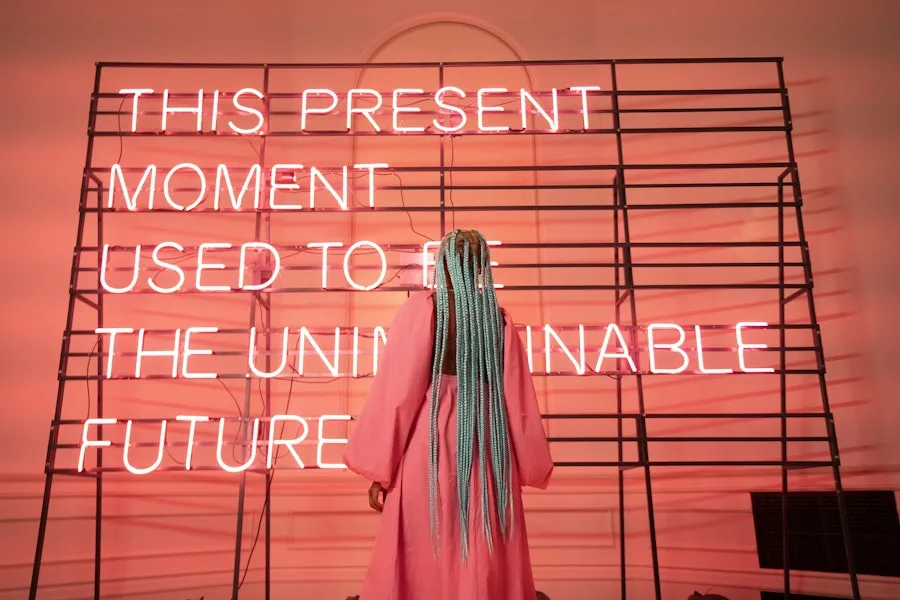 A person with long braids stands facing a glowing neon text installation in a gallery space.