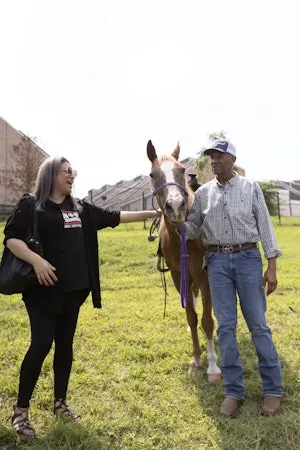 A woman and a man stand beside a horse in a grassy urban setting, smiling and interacting with the animal.