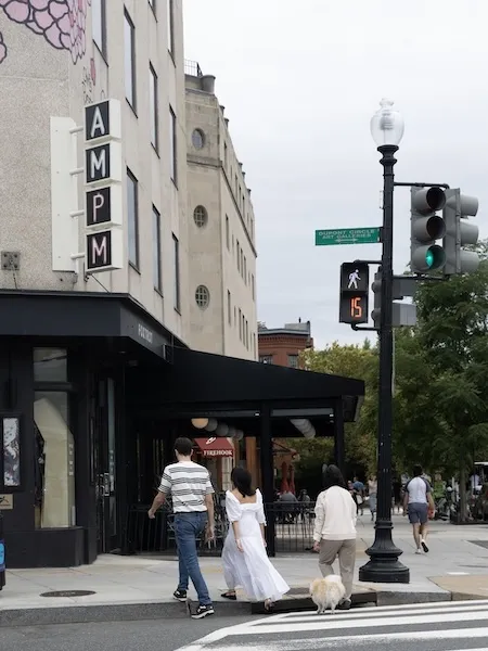 A person in a white dress crosses city street with companions near an Art Deco hotel entrance