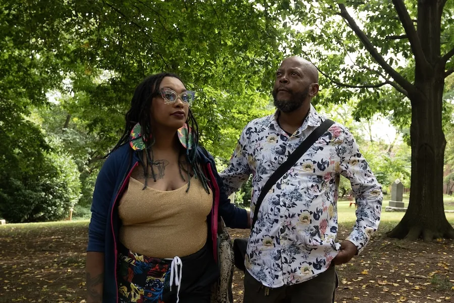 Two people in a leafy park, one holding a phone, both wearing patterned outfits.