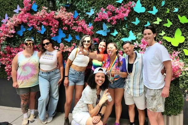 Group of people posing for a photo during Capital Pride’s Crack of Noon Brunch, standing in front of a colorful floral wall decorated with bright butterfly cutouts and rainbow-themed attire.