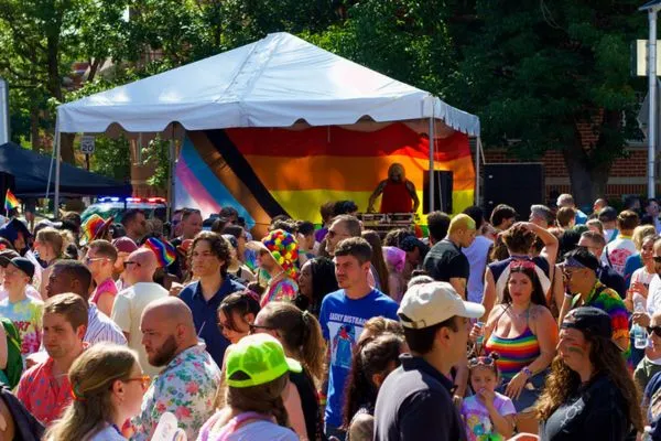 Crowds celebrating at the 17th Street Pride Block Party, gathered around a DJ booth beneath a rainbow Pride flag backdrop, with attendees wearing colorful outfits and accessories.