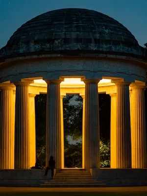 Illuminated domed pavilion with columns at dusk, warm yellow lights and a person seated inside