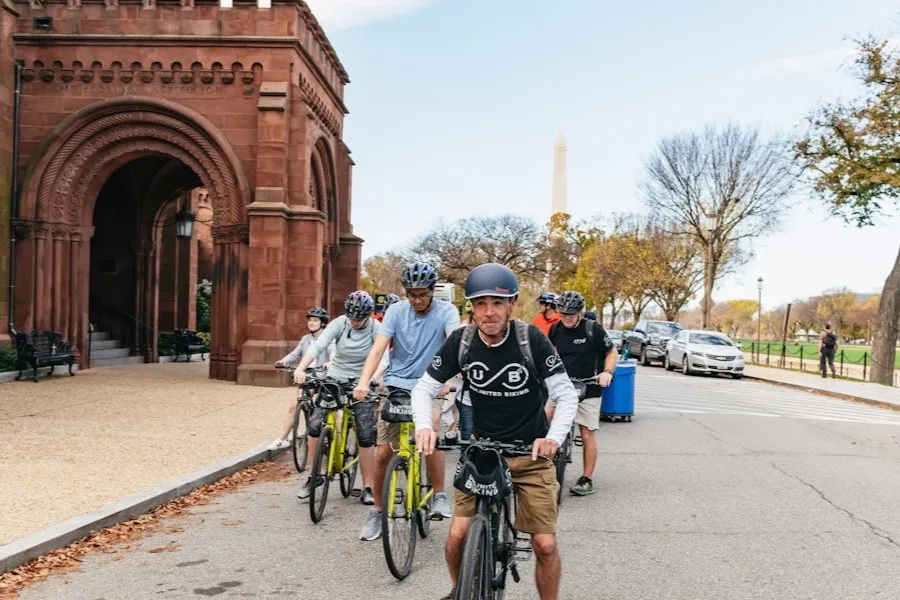 A group of cyclists rides past the Smithsonian Castle on the National Mall, with the Washington Monument visible in the background.