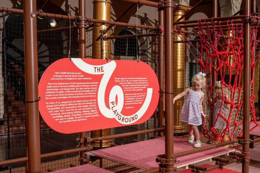 Child exploring a rope climbing structure beside an introductory sign at the National Building Museum’s PLAYGROUND exhibition.
