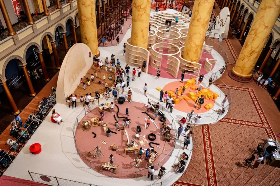 Overhead view of the National Building Museum’s PLAYGROUND exhibition, featuring hands-on building stations, a sand play area, foam blocks and a wooden maze.