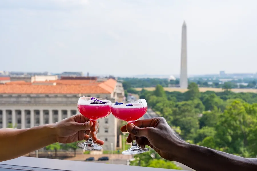 Two hands toast red cocktails with the Washington Monument in the distance