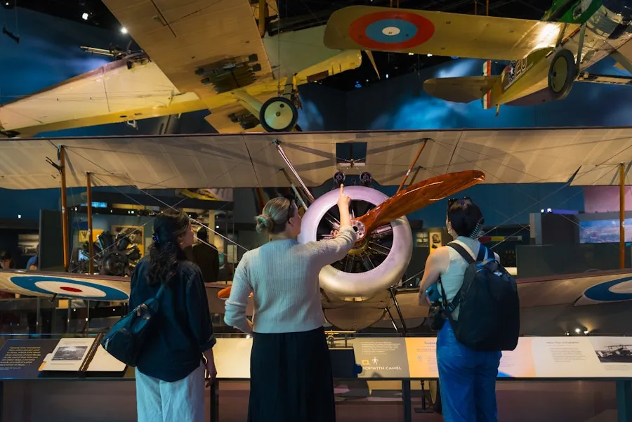 Three visitors explore a museum exhibit featuring historic aircraft, with one person examining the propeller of a vintage biplane.