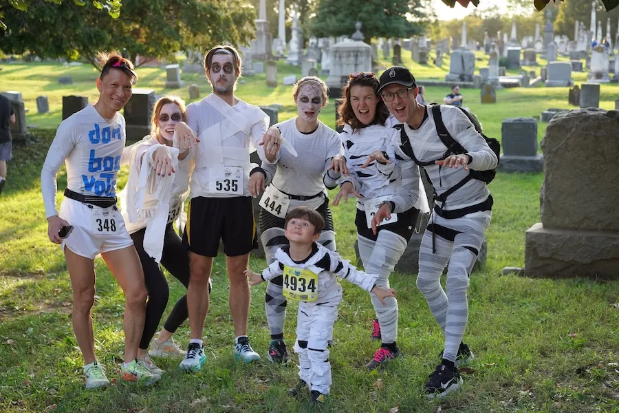 Runners posing in a cemetery dressed as mummies, one adult holding a small child in race bibs