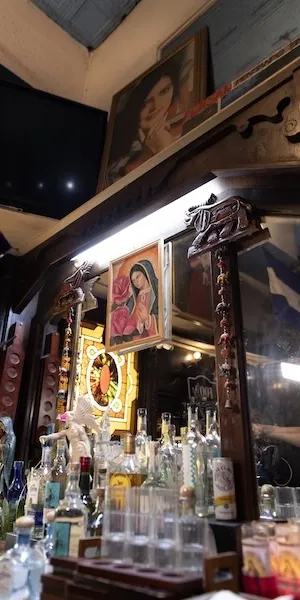 Bar shelf with liquor bottles and glassware under warm lighting, religious icons on wall