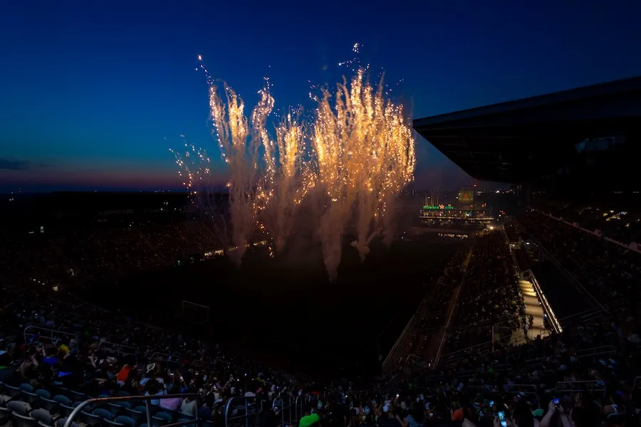 Photo of fireworks bursting above a stadium crowd at dusk