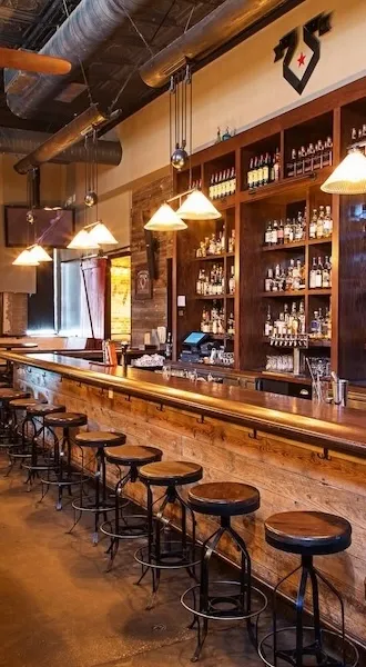 Bar interior with long wooden counter, stools and backlit shelves of bottles.