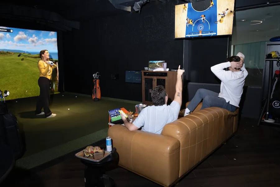 Person swings at indoor golf simulator as another lounges on tan sofa while watching sports.