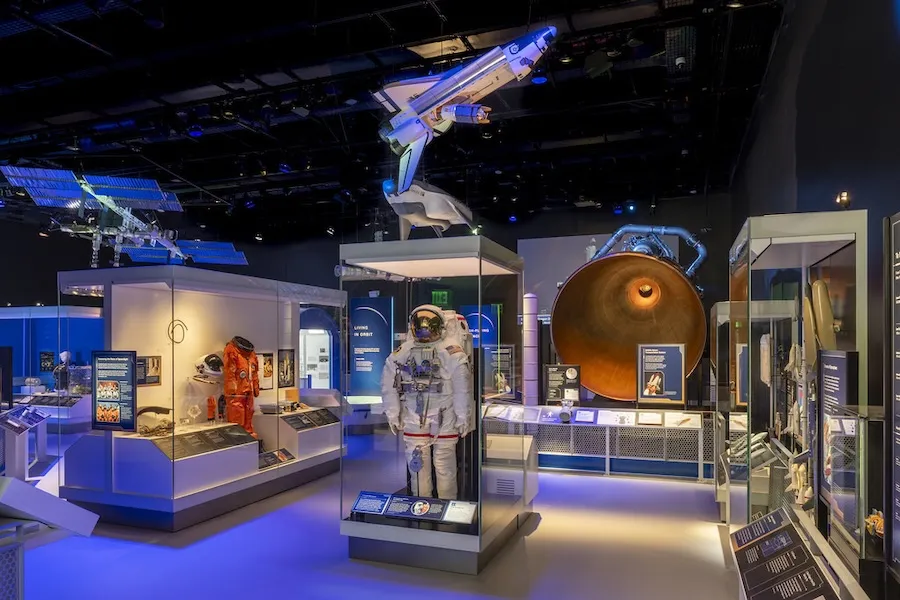 Astronaut suit on display amid spacecraft exhibits in blue-lit museum gallery
