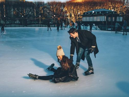 Um rinque de patinação no gelo com uma mulher que caiu, mas está rindo, e um homem rindo e ajudando-a a se levantar.